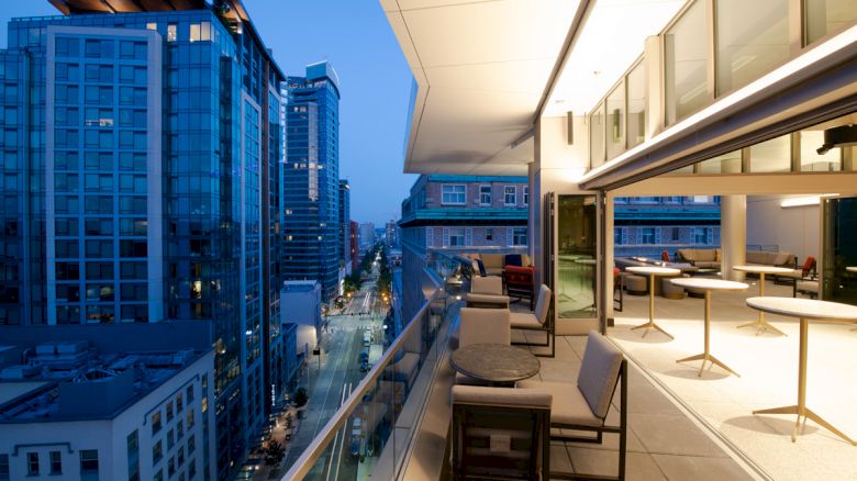 A modern balcony with seating overlooks a cityscape with tall buildings at twilight, capturing the urban skyline and streets below the scene.