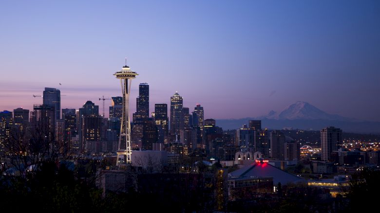 A city skyline during twilight, prominently featuring a distinct space needle structure and Mount Rainier visible in the background, under a clear sky.