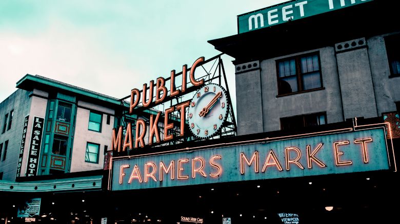 The image shows a vibrant public market with neon signs for "Public Market" and "Farmers Market," along with a clock, set against buildings.