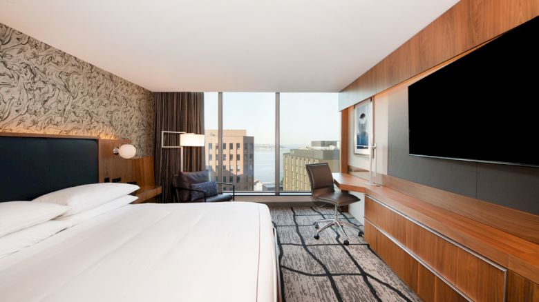 A modern hotel room features a large bed, wall-mounted TV, desk, chair, and large window with a view of city buildings and water.