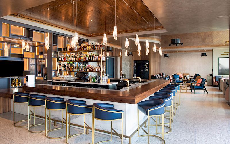 The image shows a modern bar with a wooden countertop, blue cushioned stools, and hanging lights. There are shelves filled with bottles.