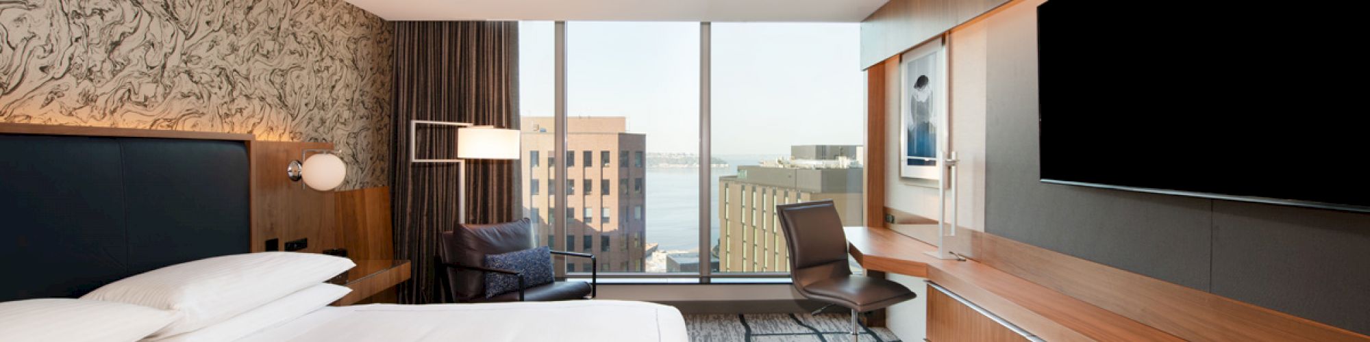 A modern hotel room features a large bed, a wall-mounted TV, a desk with a chair, and a view of buildings and water through a large window.