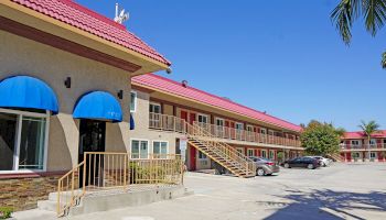 The image shows a motel with a red roof, blue awnings, and an external staircase leading to the second floor. Some cars are parked nearby in the lot.