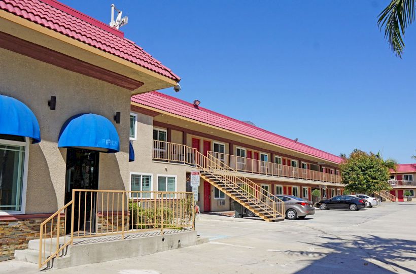 The image shows a motel with a red roof, blue awnings, and an external staircase leading to the second floor. Some cars are parked nearby in the lot.