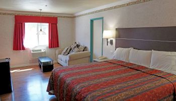 The image shows a hotel room with a large bed, a sofa, a window with red curtains, and a small table under a hanging lamp.