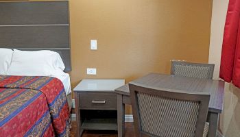 The image shows a small hotel room with a bed, a nightstand, a table, and two chairs. The wall and curtains are brown and red.