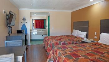 A hotel room with two double beds, wall-mounted TV, desk, and chair. The room includes an iron and board, and a visible bathroom vanity area.