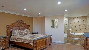 A bedroom with a large wooden bedframe, matching bedside tables, and a bathroom featuring a jacuzzi tub and beige tiled walls.