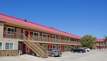 A two-story motel with red roofs, a beige exterior, and an outdoor staircase. Several cars are parked in the lot beside the trees and bushes.