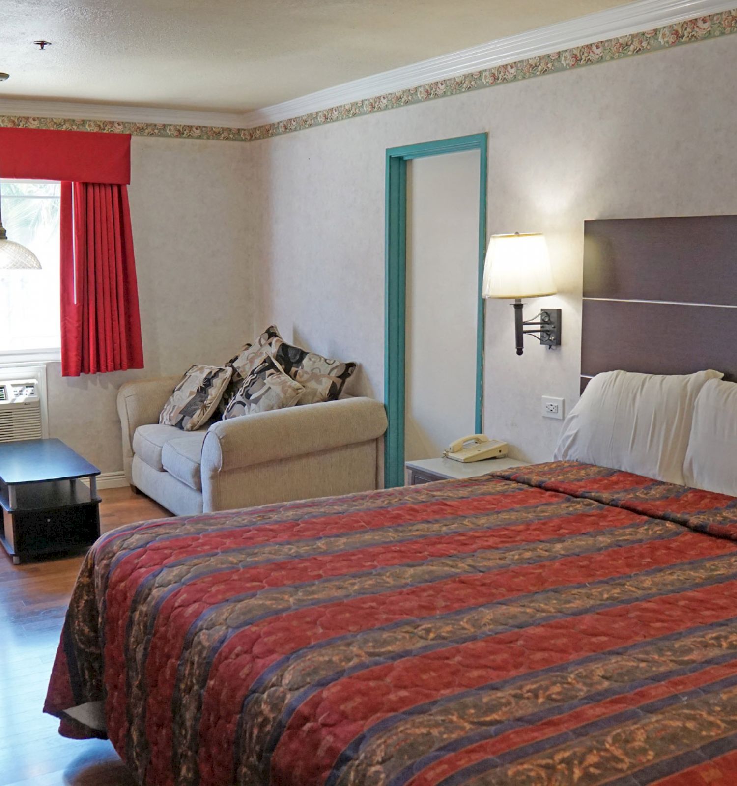 A hotel room with a king-size bed, red and orange bedspread, sofa, coffee table, lamp, window with red curtains, and wooden floor.