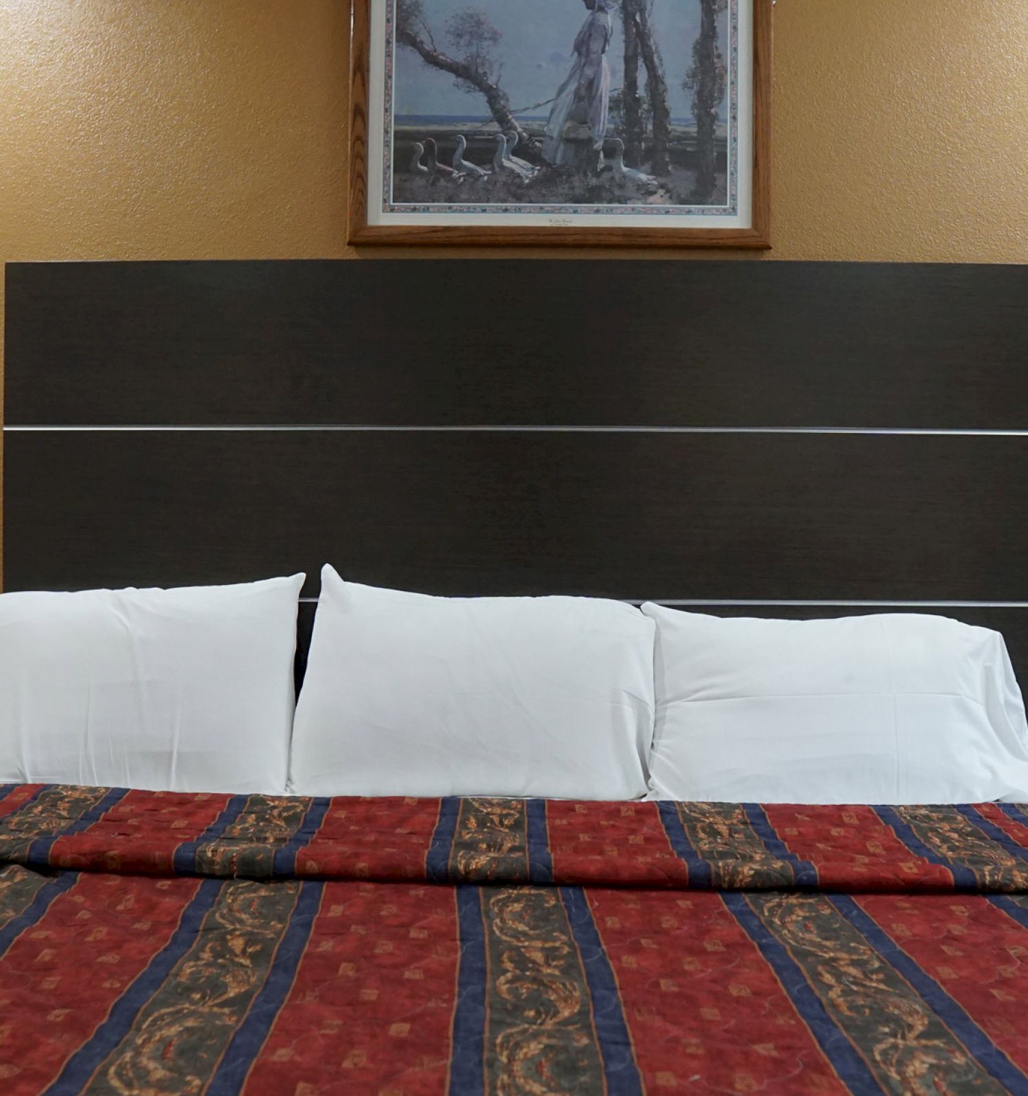 A bed with a dark headboard, white pillows, and a colorful striped blanket; a framed picture hangs on the wall above the bed.