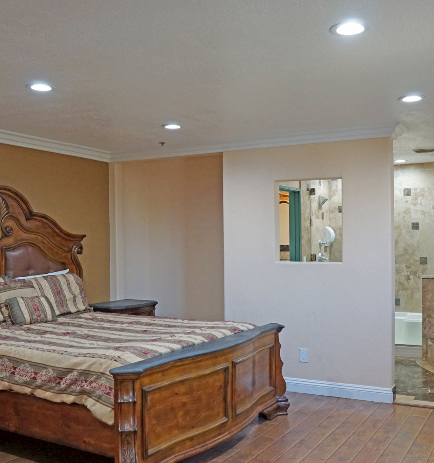 A bedroom featuring a large ornate bed with striped bedding, wooden furniture, and an open view into a tiled bathroom with a bathtub.