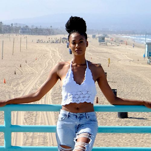 A woman leans on a turquoise railing at a beach, wearing a white top and ripped jeans, with a lifeguard tower and ocean in the background.