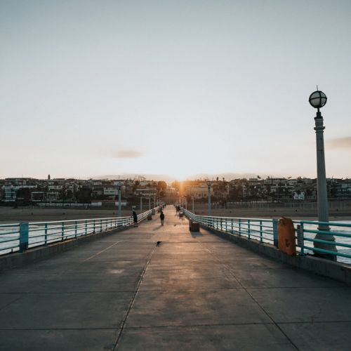 A sunset view of a pier with railings and lamp posts, leading towards the ocean with people walking and buildings visible in the background.