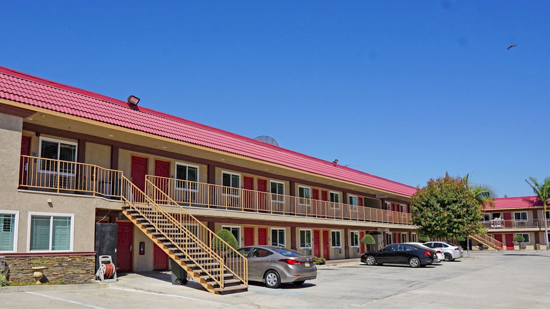 A two-story motel with a red roof, an external staircase, and parked cars in the lot. Clear blue sky and some trees in the background.