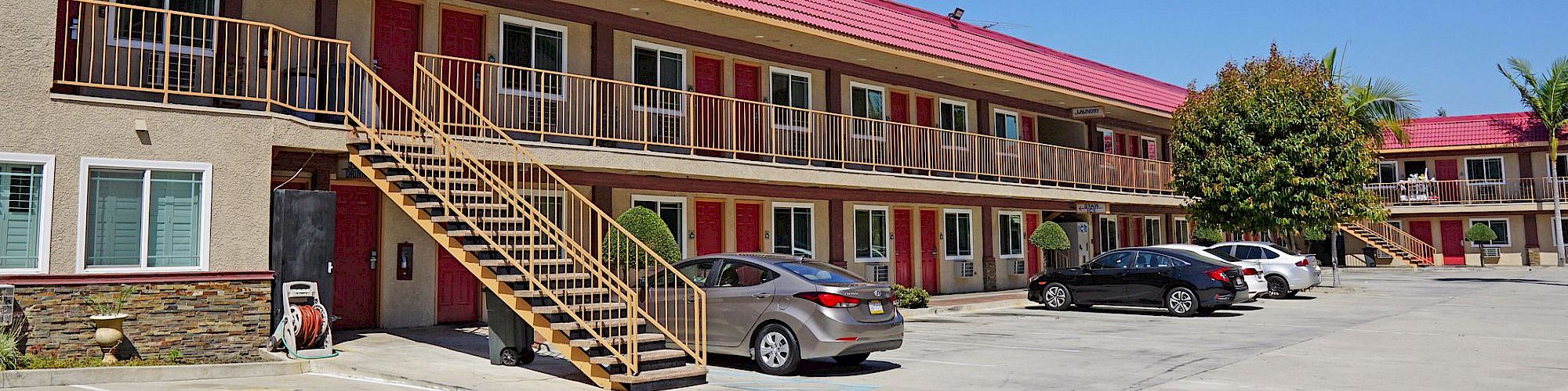 A two-story motel with a red roof, an external staircase, and parked cars in the lot. Clear blue sky and some trees in the background.